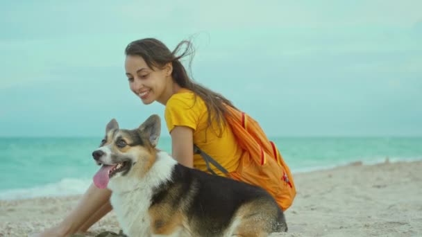 Attractive young woman smiling and spending time together with her pet cute corgi dog outdoors at sandy sea beach at sunset. — Stock Video