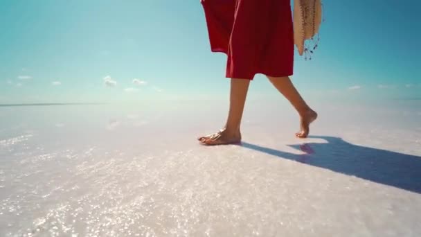 Vista lateral piernas de mujer en falda roja caminando en la playa blanca sal, sosteniendo sombrero de paja — Vídeos de Stock