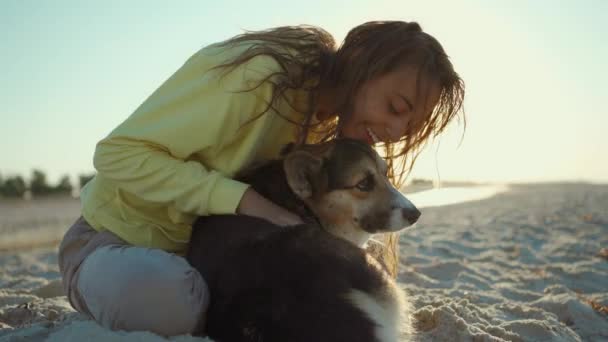 Beautiful happy and affectionate laughing girl in yellow hoodie at summer beach with cute pet Corgi dog, — Stock Video