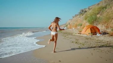 Yavaş çekim, mutlu fitness kızı, bikinili, yüksek dikenli kil sahilde kumsalda koşuyor..