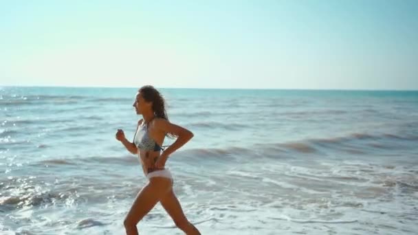 Feliz joven mujer impresionante con hermoso cuerpo delgado se dedica a la gimnasia en el mar al amanecer. Hace una carrera a lo largo de la costa. — Vídeos de Stock