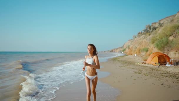 Joven mujer impresionante en bikini con hermoso cuerpo delgado se dedica a la gimnasia en el mar al amanecer. chica hace una carrera a lo largo de salvaje mar vacío costa con tienda. — Vídeos de Stock