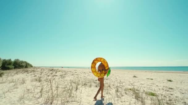 Junge schlanke, braun gebrannte Frau, die am leeren Strand mit einer riesigen aufblasbaren Ananas spaziert. Mädchen genießt Sommerurlaub am tropischen Strand — Stockvideo