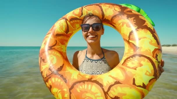 Retrato de feliz joven sonriente en gafas de sol con piña inflable de pie en agua de mar y riendo. — Vídeos de Stock