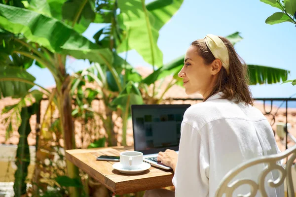 Nuori nainen istuu terassilla aamulla loman aikana, kannettavan tietokoneen avulla — kuvapankkivalokuva