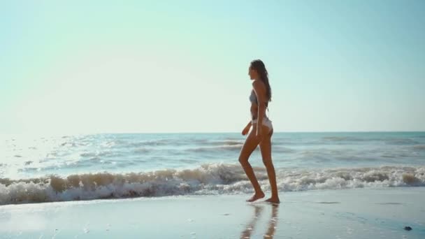 Imágenes en cámara lenta de la alegre chica de fitness feliz en bikini camina a lo largo de la playa de mar con olas — Vídeo de stock