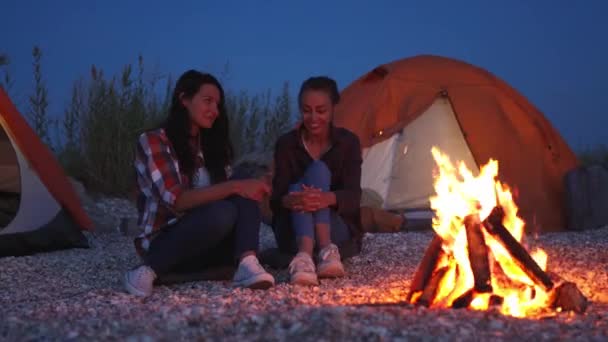 Две красивые девушки сидят у костра рядом с открытой оранжевой палаткой — стоковое видео
