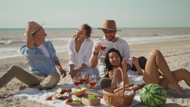 Vier glückliche Millennial-Freunde picknicken am Sandstrand am sonnigen Tag am nächsten winkenden Meer — Stockvideo