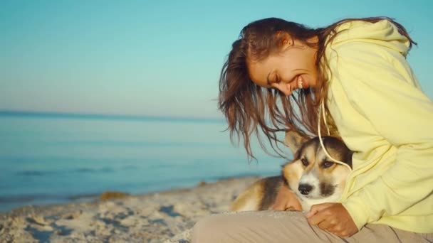 Candid risa chica latina en sudadera con capucha amarilla en la playa de verano con lindo perro Corgi mascota, sentado en el mar frontal con la luz del sol de la mañana. — Vídeo de stock