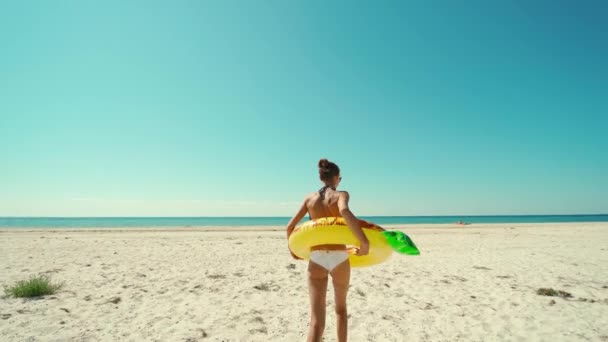 Camera volgen voor jonge vrouw met perfect slank gebruind lichaam en sexy billen lopen op leeg strand naar water houden reusachtige opblaasbare ananas. — Stockvideo