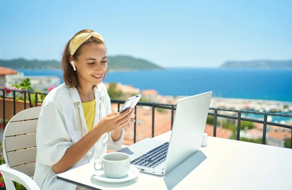 En kvinne i trådløs hodetelefon som ser på smarttelefon mens hun sitter på balkongen med Seaview på ferie – stockfoto