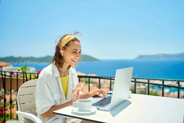 Kaunis valkoihoinen nainen langattomissa kuulokkeissa puhuvat videopuhelun tietokoneella istuessaan parvekkeella merinäköalalla lomakeskuksessa. — kuvapankkivalokuva