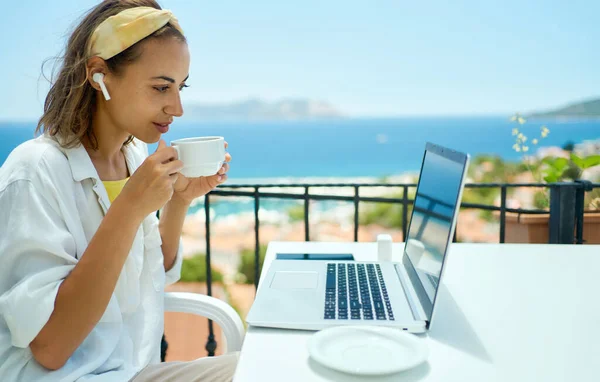 Nuori sekarotuinen nainen langattomissa korvakoruissa juomassa kahvia ja katselemassa kannettavan tietokoneen näyttöä istuessaan parvekkeella lomahotellissa — kuvapankkivalokuva