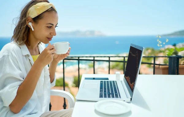 Nainen juo kahvia ja katselee kannettavaa tietokonetta istuessaan terassilla meren rannalla — kuvapankkivalokuva
