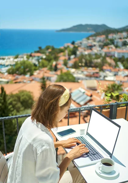 Nainen istuu hotellin parvekkeella kauniilla merinäköalalla lomakohteessa, työskentelee kannettavalla tietokoneella — kuvapankkivalokuva