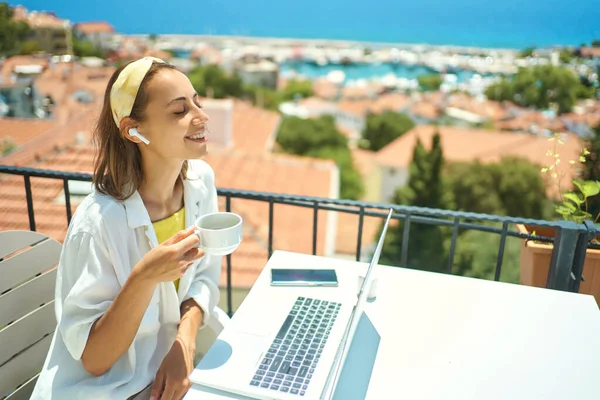 Pleased woman enjoying coffee while sitting on terrace on sea resort, using laptop computer for remote work — kuvapankkivalokuva