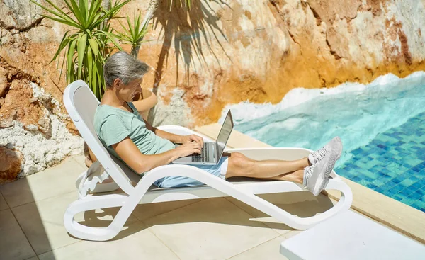 En mann som slapper av ved bassenget i ferien, skriver på tastatur, chatter med venner – stockfoto