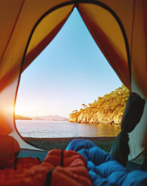 Sett fra innsiden av et telt på en vill strand ved soloppgang og beina til to personer i soveposer – stockfoto