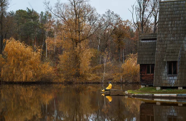 Fille en vêtements jaunes assis sur la jetée, la surface de l'eau calme du lac reflète le feuillage orange et les arbres — Photo