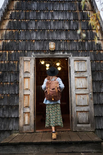 Femme touristique élégante dans un chapeau avec sac à dos à venir dans une vieille cabane en bois de style vintage — Photo