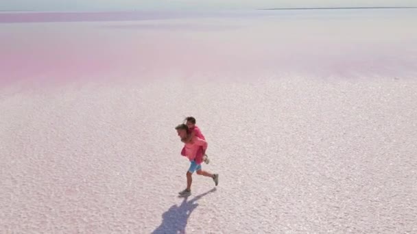 Vue aérienne de drone de couple heureux en amour sur des vacances d'été en plein air. Joyeuse fille piggybackking sur copain avoir du plaisir à marcher sur la rive de beau lac rose coloré — Video