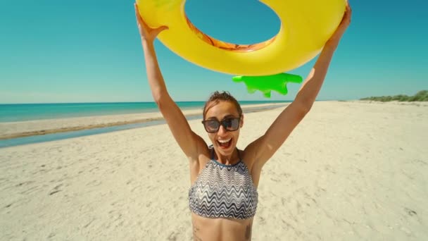 Усміхнена дівчина на пляжі сонячного океану дивиться на камеру через жовте надувне кільце зі смішним обличчям — стокове відео