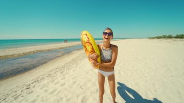 Neşeli, genç, güzel, bin yıllık bir kadın boş kumlu sahilde elinde sarı şişme ananas arabasıyla yazın.