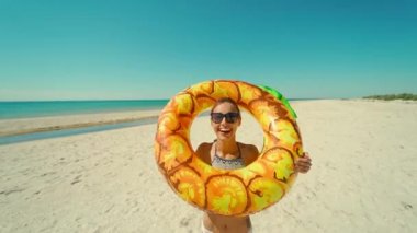 Güneşli okyanus sahilinde gülümseyen kız komik suratlı sarı şişirilebilir halkadan kameraya bakıyor.