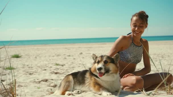 Gelukkige liefdevolle en tedere gebruinde vrouw in bikini liggend op zand aan het zonnige zeestrand met haar huisdier, schattige welsh corgi hond. meisje strelen en kussen haar hond — Stockvideo