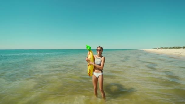 Портрет счастливой улыбающейся молодой женщины в солнечных очках с надувным ананасом, стоящим в морской воде и смеющейся. — стоковое видео