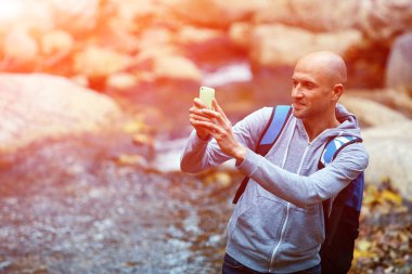 uzun yürüyüşe çıkan kimse adam dere kıyısında ormanda gülümseyen ve Smartphone'da fotoğraf yapma