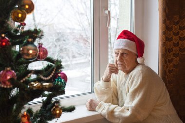 Noel Baba şapkalı yaşlı beyaz kadın pencerenin kenarında oturur ve ne yazık ki Noel ağacına bakar. Kavram: Noel arifesinde yalnız yaşlı insanlar. Portre. Yakın plan..