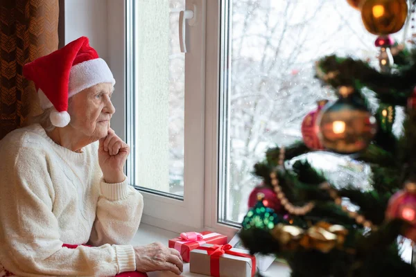 サンタの帽子をかぶった高齢の白人女性が窓のそばに座っている クリスマスの前夜 贈り物 クリスマスツリー コンセプト 魔法の休日 孤独を待っている 肖像画 — ストック写真