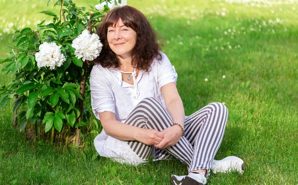 Portrait of  beautiful woman of forty-five years old with brown hair, sitting on green lawn in sun and near flower bush of white peonies. Summer weekend. Focus on woman.