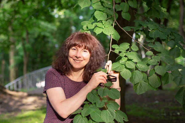 Pretty woman of forty-five with brown hair looks at camera and holds a linden branch in her hands. Portrait of positive smiling woman. Sunlight. Summer evening in the park