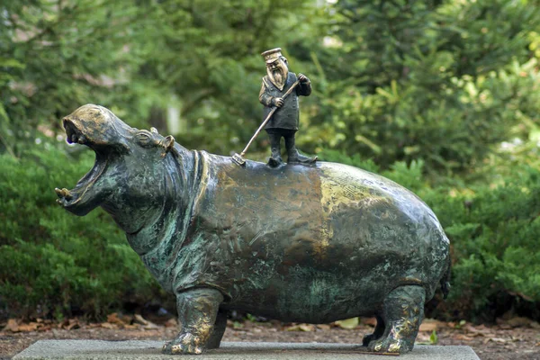 A metal statue of a hippo, which is also cleaned by a tiny metal man. This monument stands in the Wrocaw Zoo surrounded by greenery.