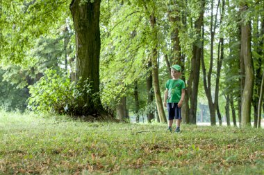 Beyzbol şapkalı dalgın, sevimli bir çocuk tam önüne bakıyor. Arka planda, yeşil ağaçlar ve diğer bitkiler.