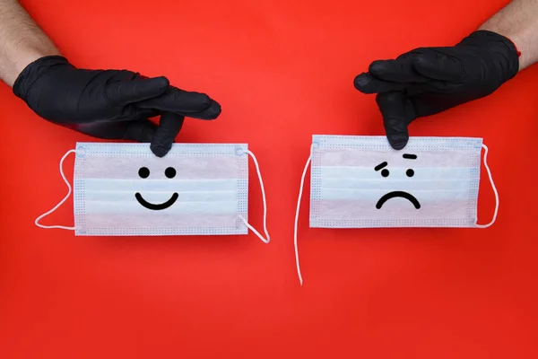 Ręce w czarnych rękawiczkach lateksowych posiadają dwie maski medyczne, na których śmieszny i smutny uśmiech — Zdjęcie stockowe