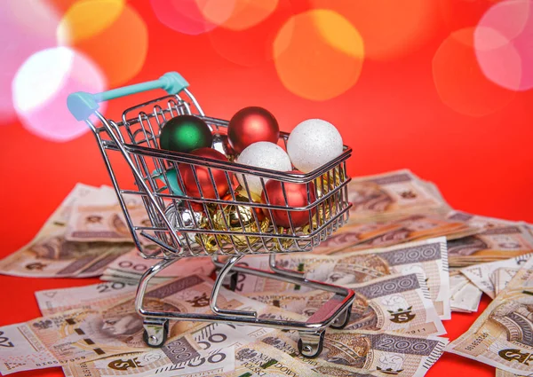 Small shopping cart full of balls from Christmas tree stands on lot of banknotes — Stock Photo, Image