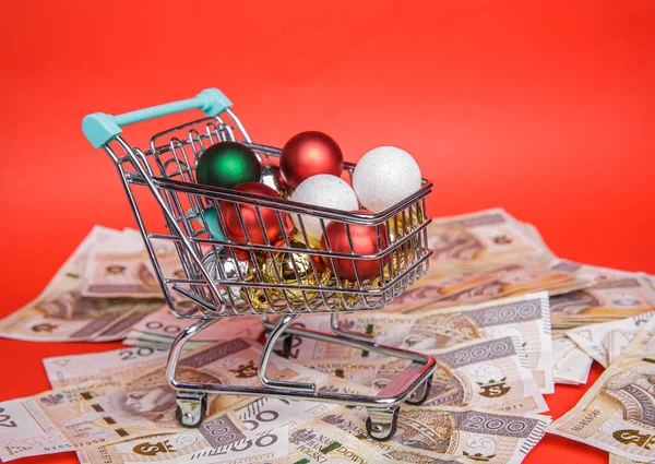 Small shopping cart full of balls from Christmas tree stands on lot of banknotes — Stock Photo, Image