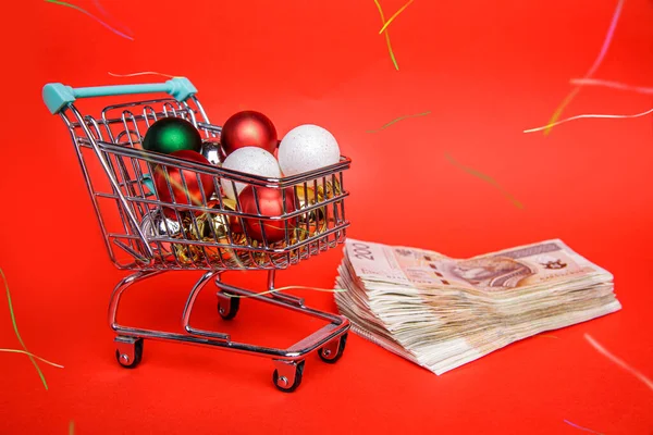 Small shopping cart full of balls from Christmas tree stands on lot of banknotes — Stock Photo, Image