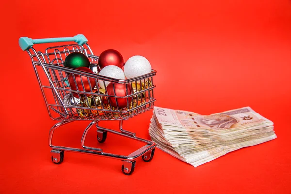Small shopping cart full of balls from Christmas tree stands on lot of banknotes — Stock Photo, Image