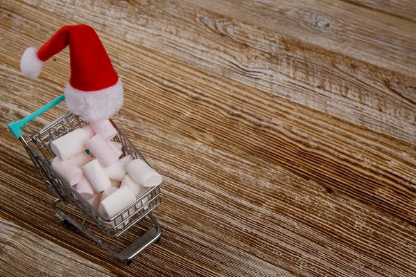 Small shopping cart filled with marshmallows and Santa hat. — Stock Photo, Image