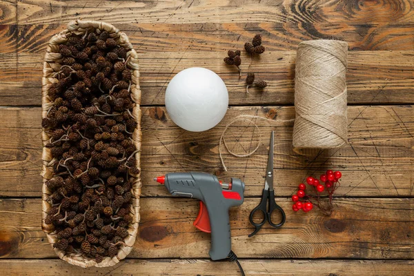 Yaratıcı Noel zanaat hobisi. El yapımı xmas koni topu yapıyorum. Talimatlar. Birinci adım: aletler, koniler, penoplast topu, ip, glu, makas. Tahta masa arkasında tatil süslemeleri. Üst görünüm