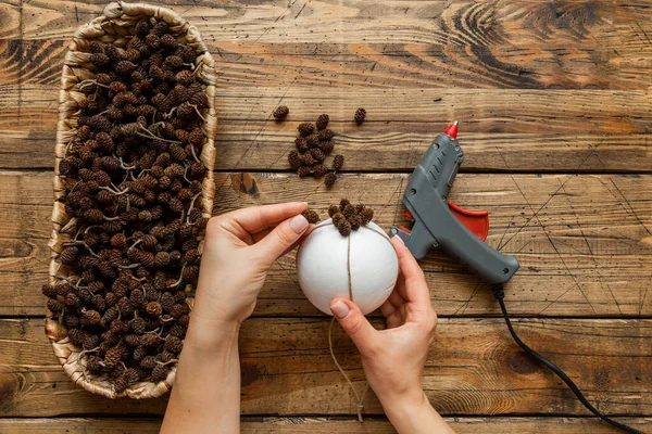 Yaratıcı Noel zanaat hobisi. El yapımı xmas koni topu yapıyorum. Talimatlar. Beşinci adım: konileri top tabanına yapıştır. Tahta masa arkasında tatil süslemeleri. Üst görünüm