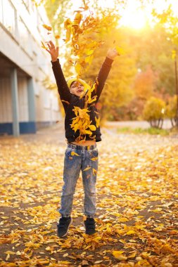 Küçük mutlu çocuk sonbahar sokağına atlayıp yaprakları fırlatıyor. Sonbahar sezonu neşeli yürümeye başlayan çocuk dikey görüntüsüyle birleştiriliyor