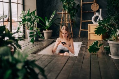 Güzel genç bir kadın şampanya içiyor ve akıllı telefona bakıyor köpük banyosunun keyfini çıkarıyor.