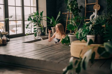 Güzel genç bir kadın şampanya içiyor ve akıllı telefona bakıyor köpük banyosunun keyfini çıkarıyor.