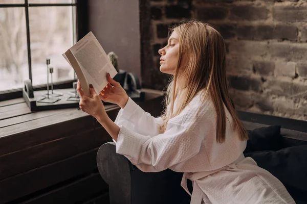Молода блондинка красива жінка в білому халаті сидить біля вікна на дивані і читає книгу . — стокове фото