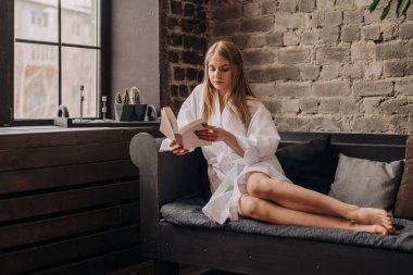 Beyaz bornozlu genç sarışın kadın kanepedeki pencerenin yanında oturuyor ve kağıt kitap okuyor..